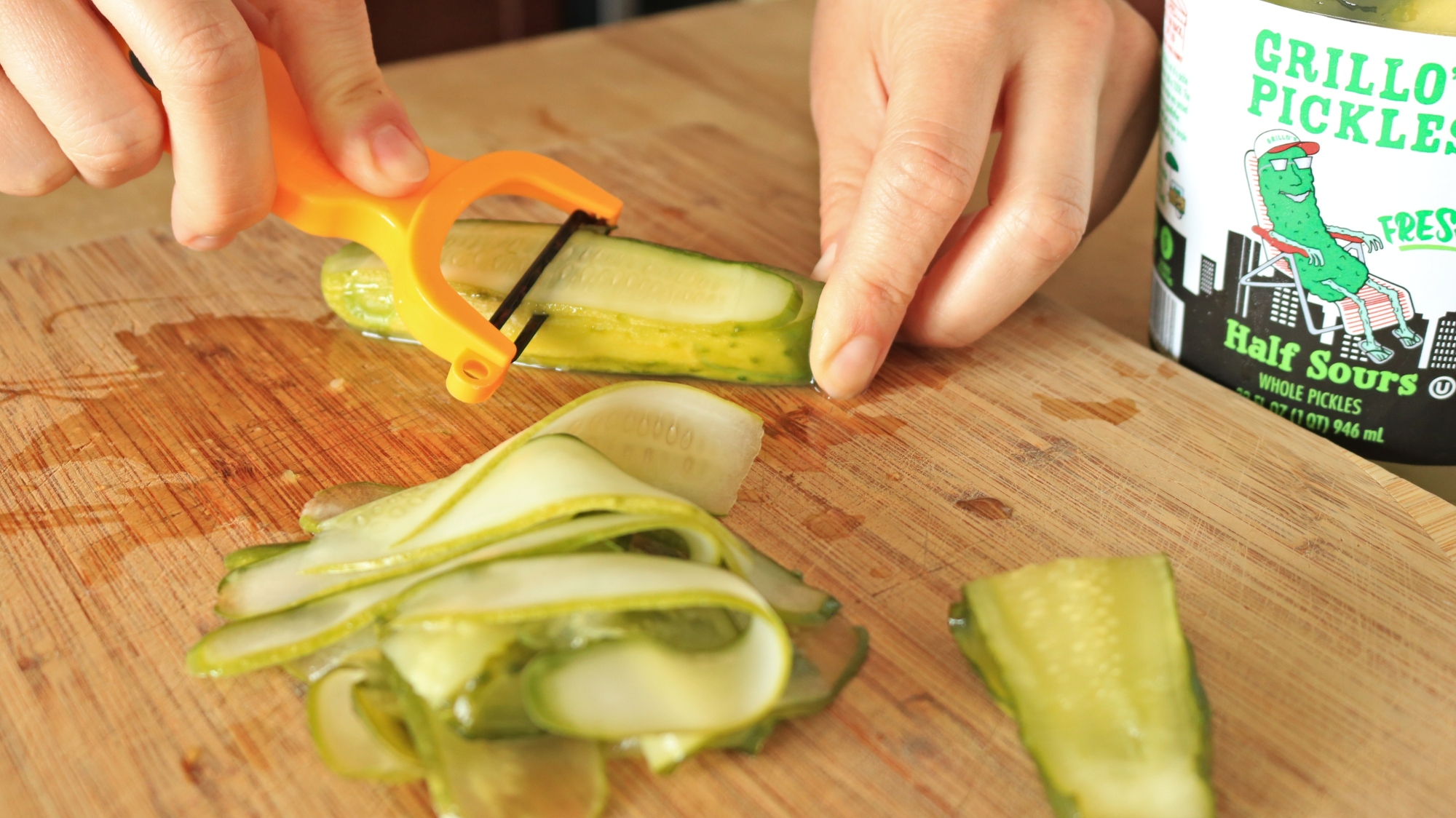 Hands using vegetable peeler to shave a pickle.